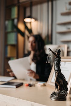 Close-up of a justice figurine on desk with blurred female lawyer in office setting.